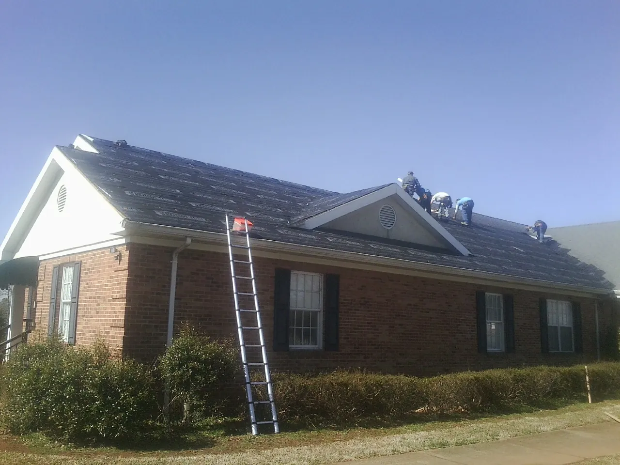 Crew tearing shingles off a brick ranch roof with a ladder set — Emergency Roof Repair work by our crew in College Station
