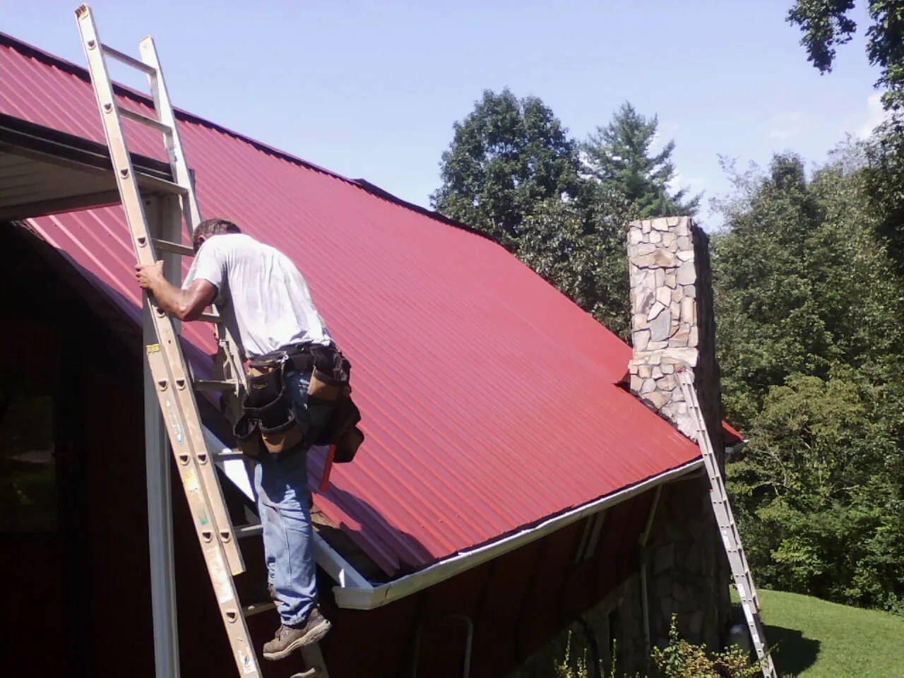 Roofer on a ladder beside a red metal roof and stone chimney — Soffit & Fascia Repair by Roofing Brothers in College Station